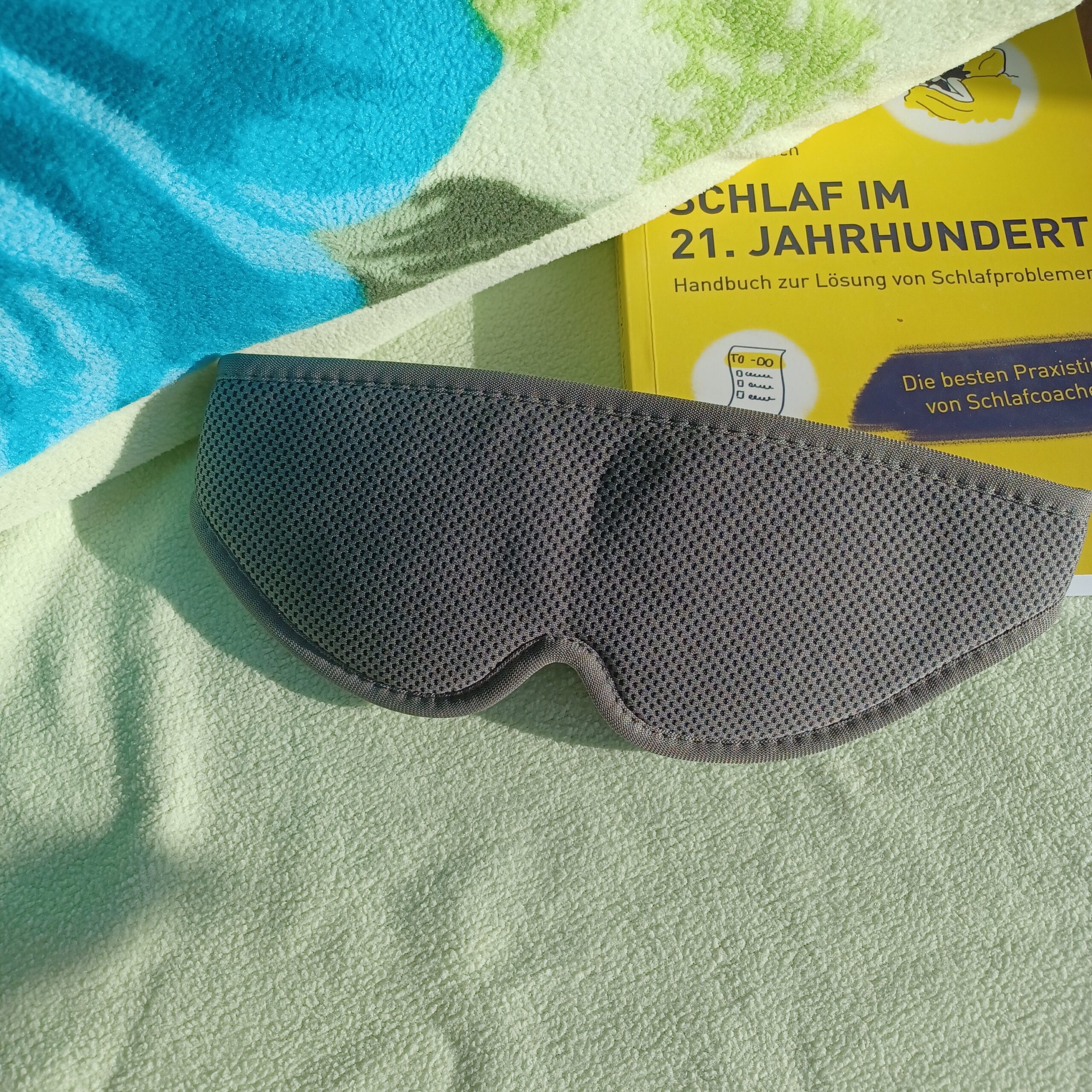 Auf einem Bett mit hellgrünem Kopfkissen liegt die sehgut Schlafbrille mit einstellbaren Augenmuscheln und dahinter ein gelbes Buch zum Thema Schlafprobleme.