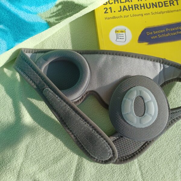 Auf einem Bett mit hellgrünem Kopfkissen liegt die sehgut Schlafbrille mit einstellbaren Augenmuscheln und dahinter ein gelbes Buch zum Thema Schlafprobleme.