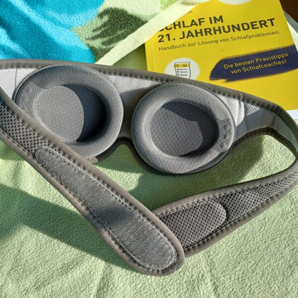 Auf einem Bett mit hellgrünem Kopfkissen liegt die sehgut Schlafbrille mit einstellbaren Augenmuscheln und dahinter ein gelbes Buch zum Thema Schlafprobleme.