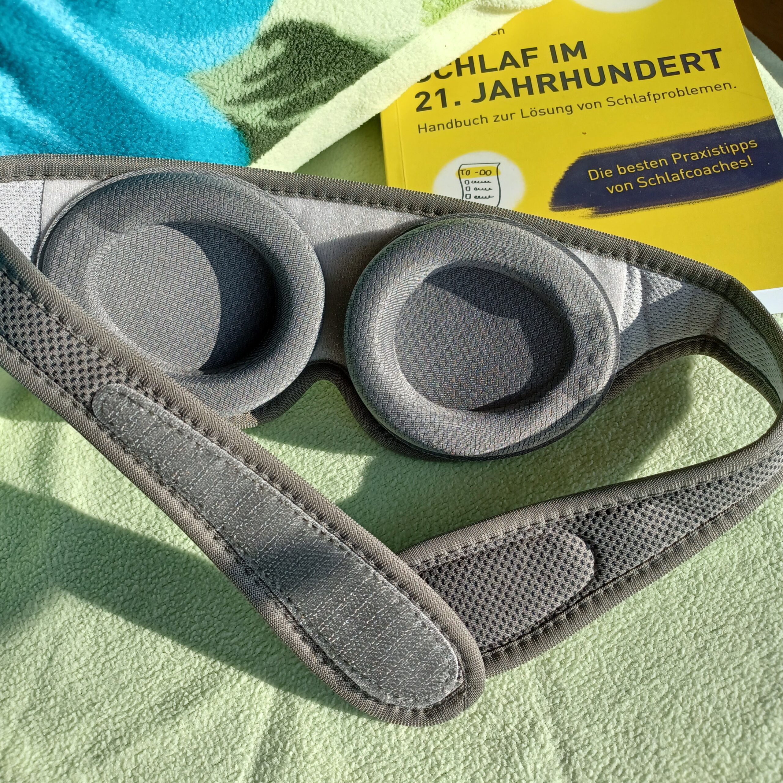 Auf einem Bett mit hellgrünem Kopfkissen liegt die sehgut Schlafbrille mit einstellbaren Augenmuscheln und dahinter ein gelbes Buch zum Thema Schlafprobleme.
