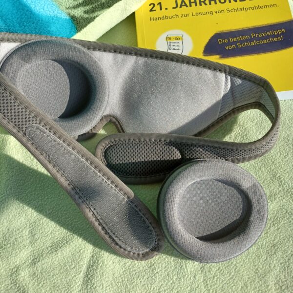 Auf einem Bett mit hellgrünem Kopfkissen liegt die sehgut Schlafbrille mit einstellbaren Augenmuscheln und dahinter ein gelbes Buch zum Thema Schlafprobleme.