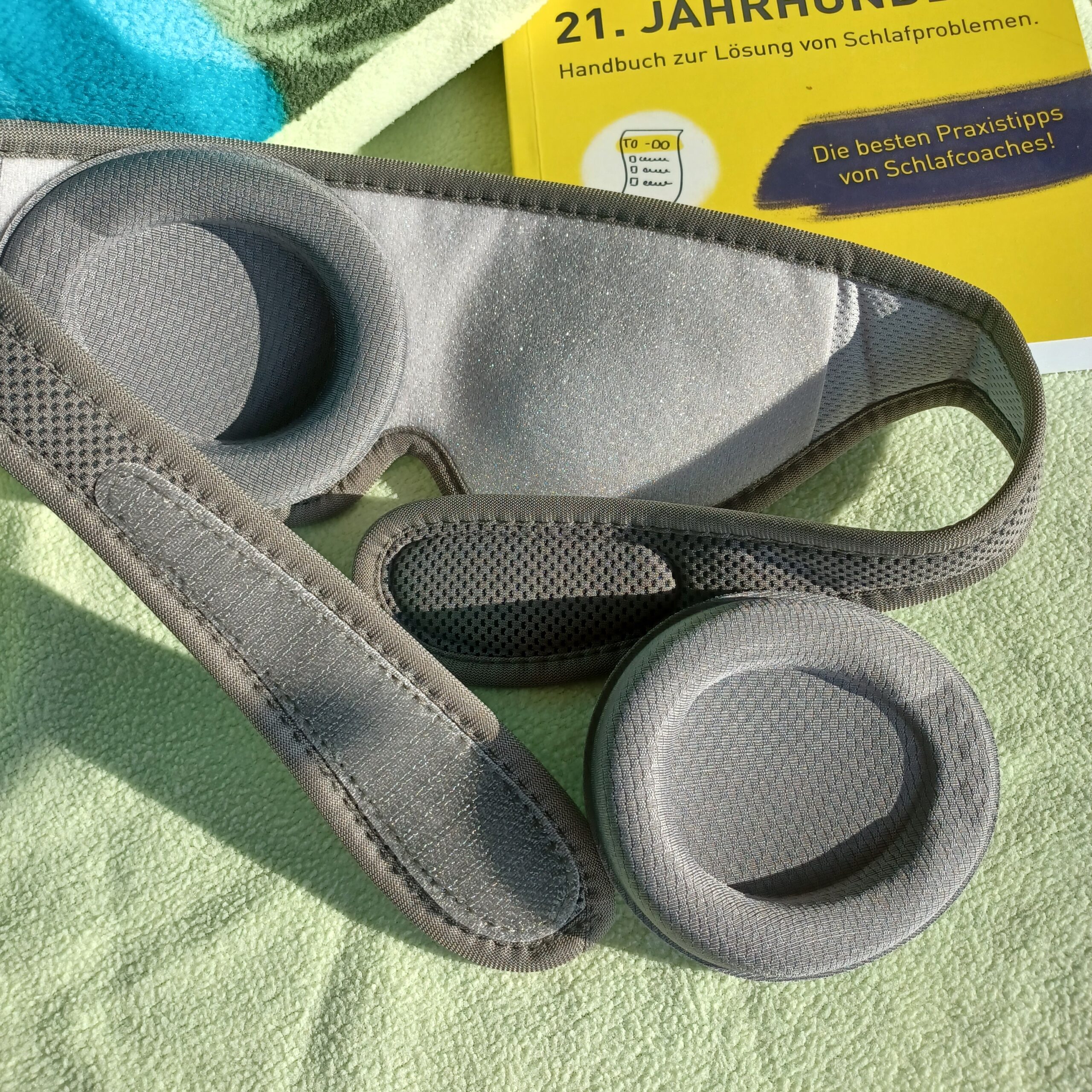 Auf einem Bett mit hellgrünem Kopfkissen liegt die sehgut Schlafbrille mit einstellbaren Augenmuscheln und dahinter ein gelbes Buch zum Thema Schlafprobleme.