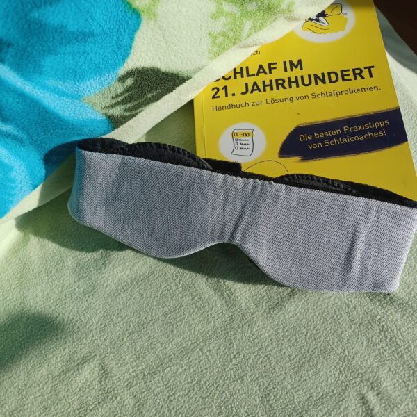 Auf einem Bett mit hellgrünem Kopfkissen liegt die sehgut Schlafbrille mit einstellbaren Augenmuscheln und dahinter ein gelbes Buch zum Thema Schlafprobleme.