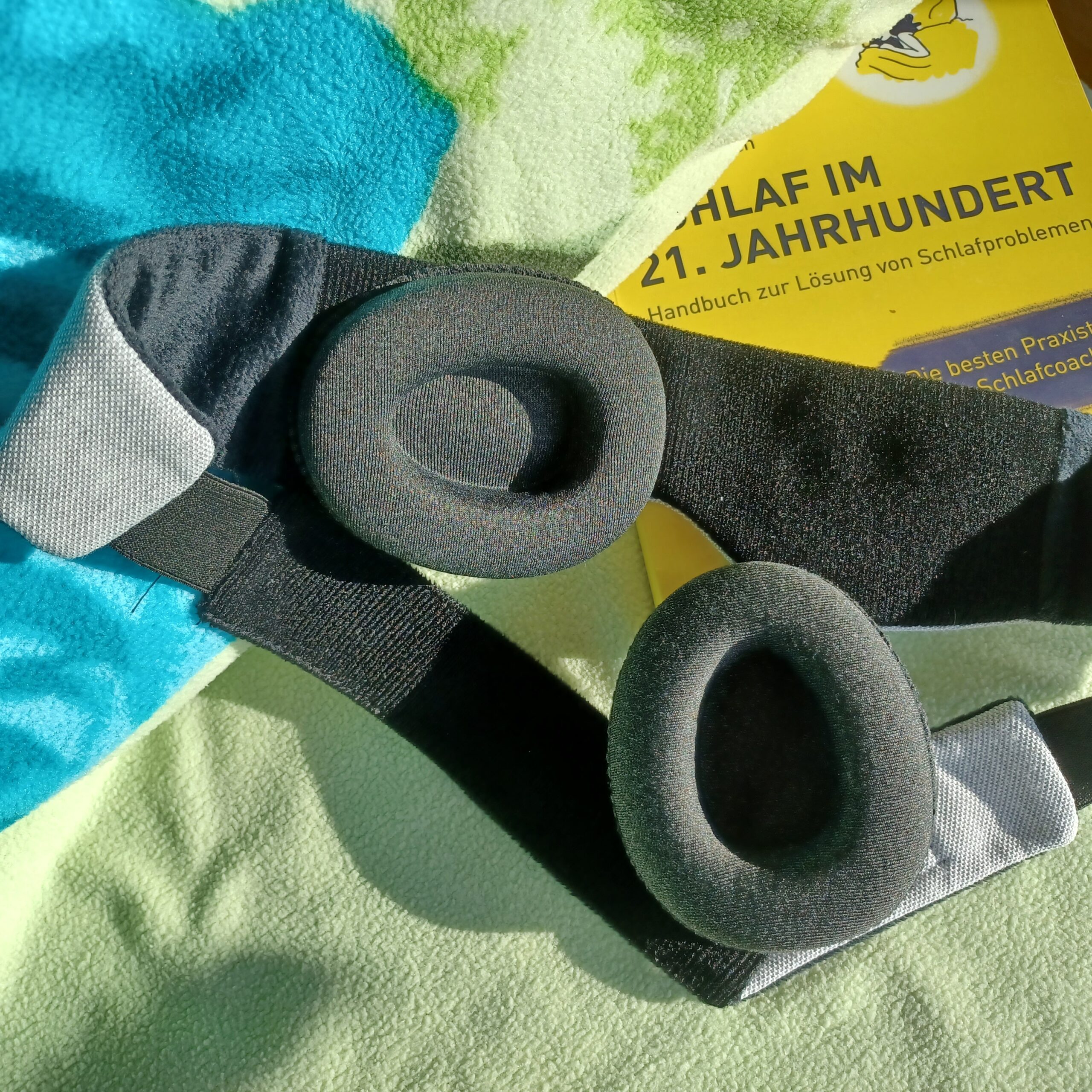 Auf einem Bett mit hellgrünem Kopfkissen liegt eine Schlafbrille mit einstellbaren Augenmuscheln von sehgut und dahinter ein gelbes Buch zum Thema Schlafprobleme.