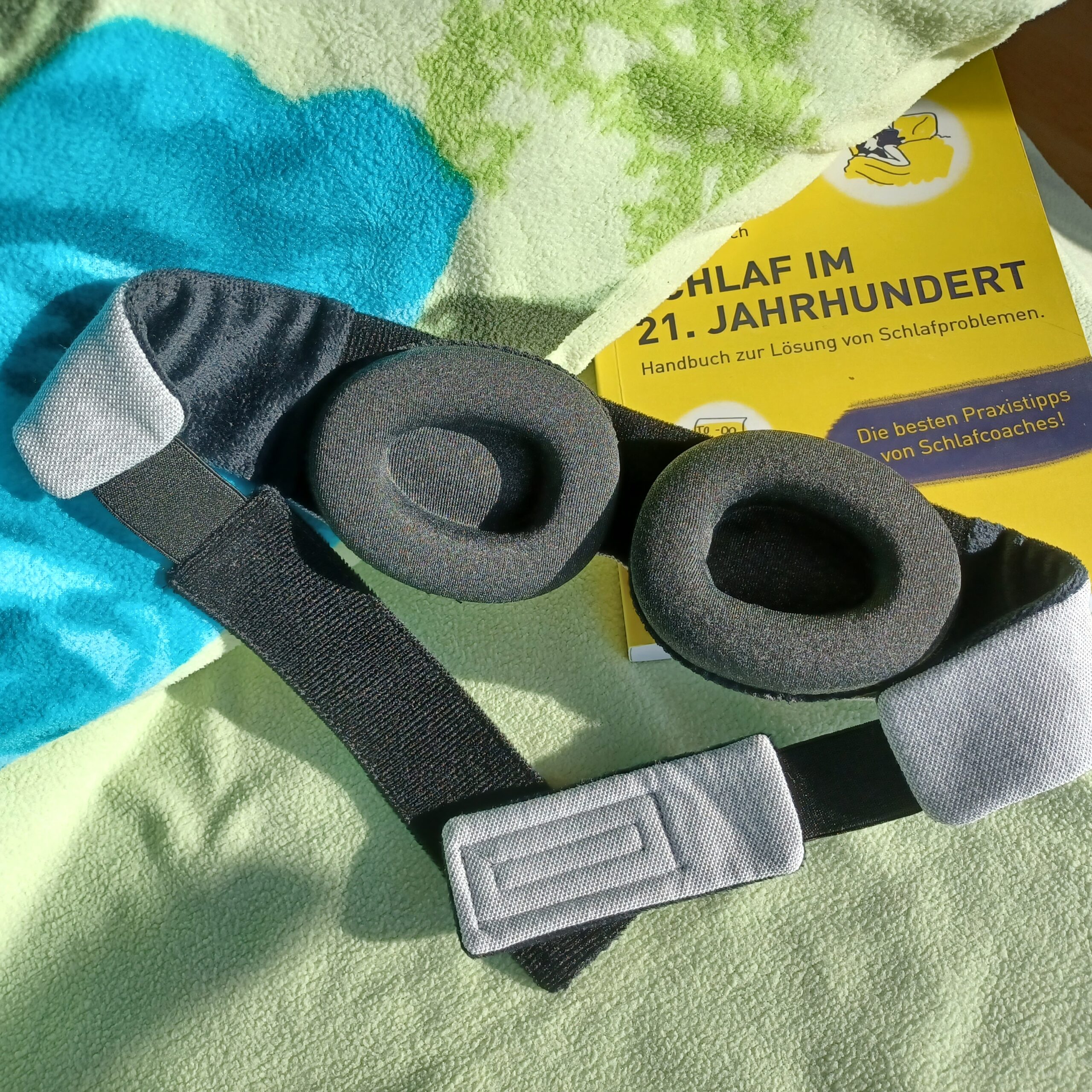 Auf einem Bett mit hellgrünem Kopfkissen liegt eine Schlafbrille mit einstellbaren Augenmuscheln von sehgut und dahinter ein gelbes Buch zum Thema Schlafprobleme.
